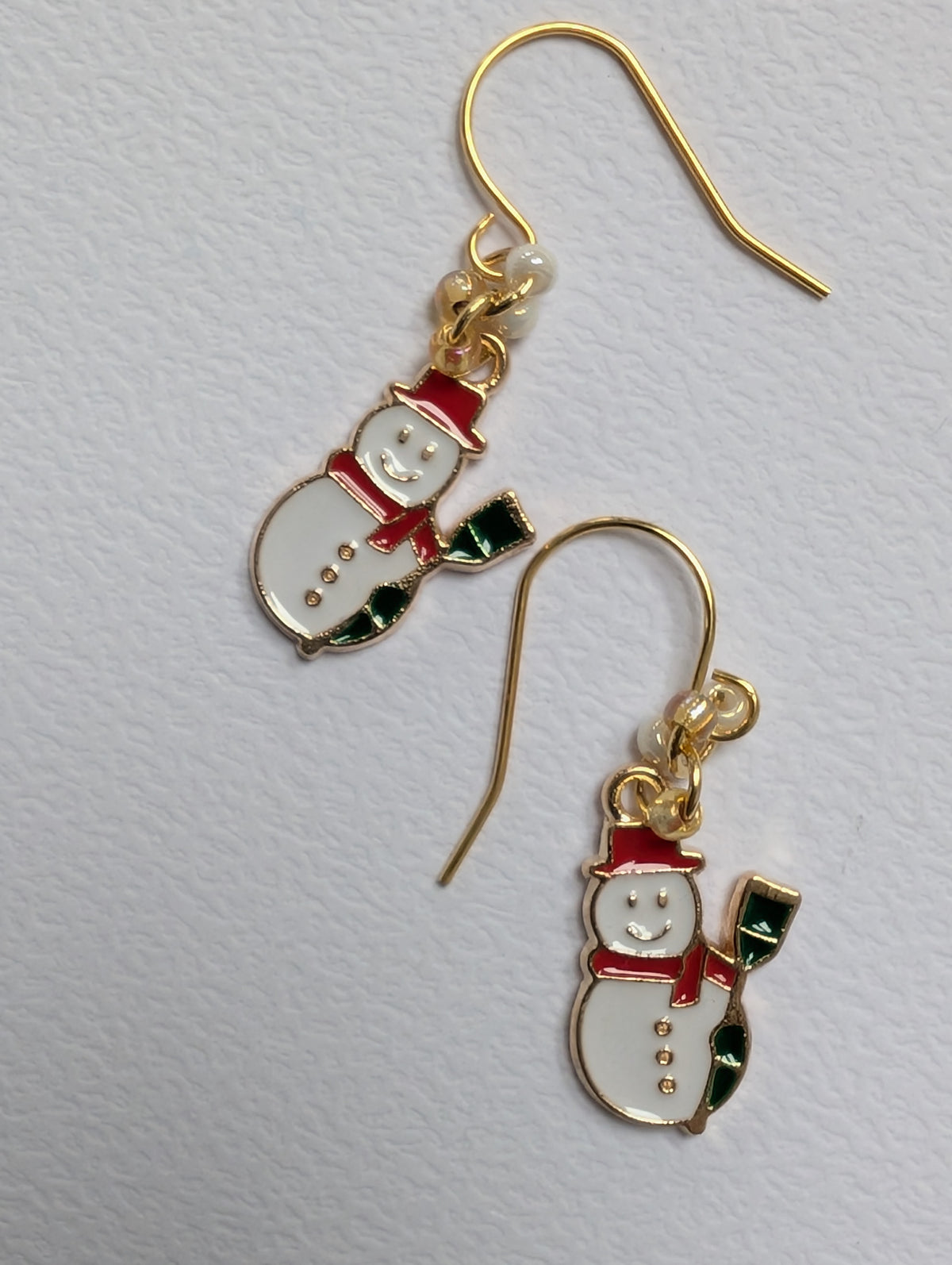 A pair of gold-tone dangle earrings featuring enamel snowman charms with white bodies, red hats and scarves, and green accents, displayed on a textured white background.