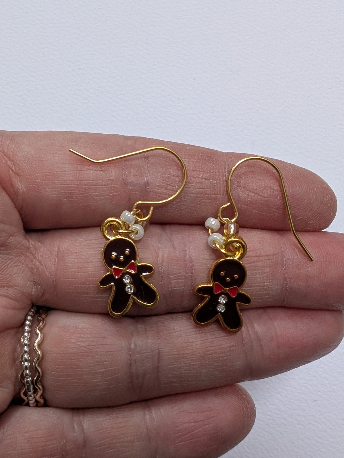 A pair of gold-tone dangle earrings with gingerbread man charms, held in a person's hand. The earrings feature dark brown and gold enamel, small white pearl-like beads, and a tiny red bow tie detail.